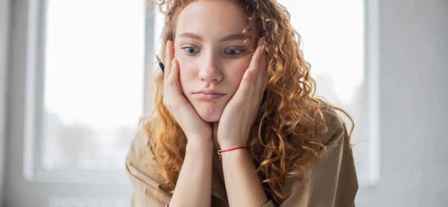 Pensive female student with hands on cheeks looking down while sitting in light room on blurred background while deciding task in frustration